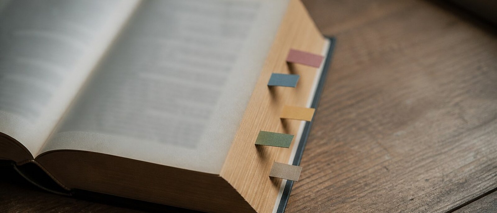 Aufgeschlagenes dickes Buch auf einem Holztisch, aus dem Seitenschnitt ragen fünf farbige Markierungsfähnchen